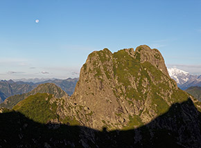 I raggi del Sole mattutino scoprono la spettacolare Cima Pedum e sulla linea d’ombra disegnano la sagoma del Bivacco di Bocchetta di Campo.